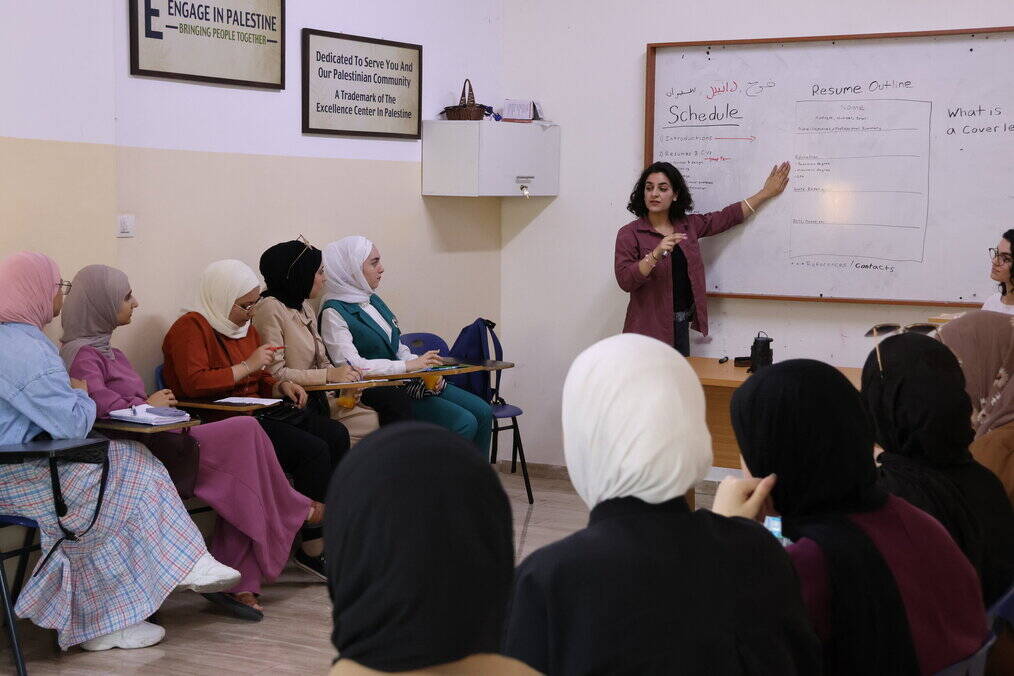A woman points at a whiteboard while adult students sitting in desks take notes.