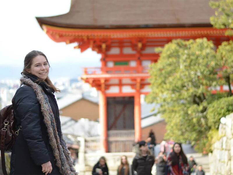 A woman wearing a a backpack poses in front of Japanese architecture.