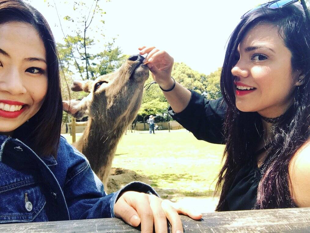 Two women smile for the camera as one feeds a deer.