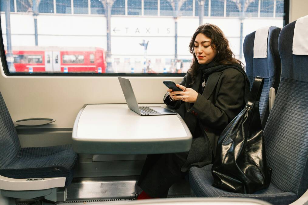 A woman on a train looks at her phone with a laptop on the table in front of her.