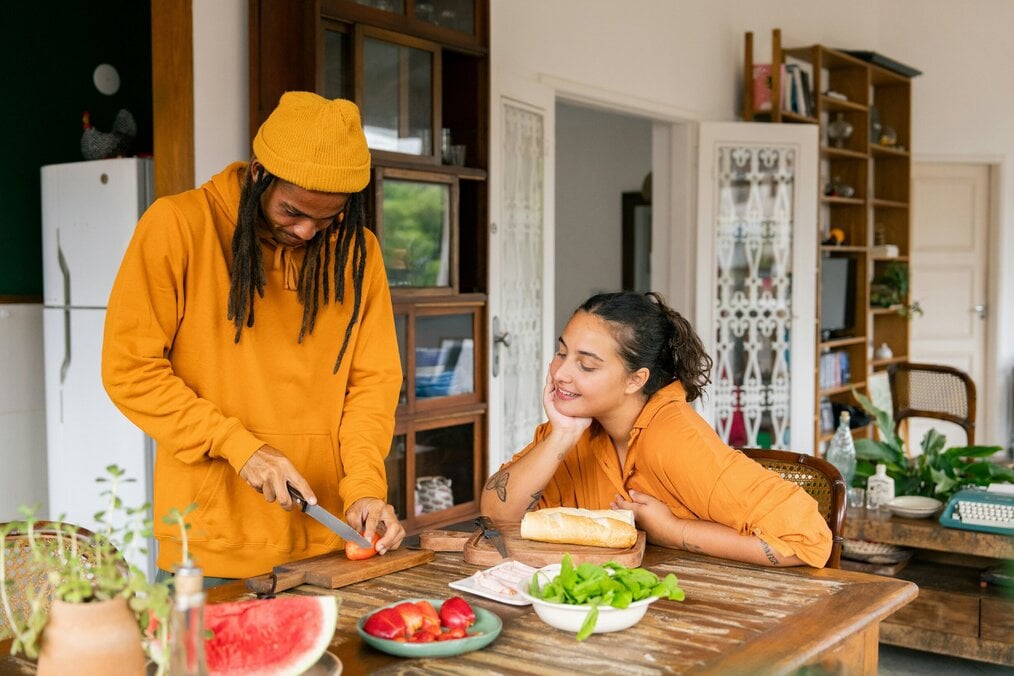 Friends cooking A man cuts a tomato while a woman sitting at the table watches.