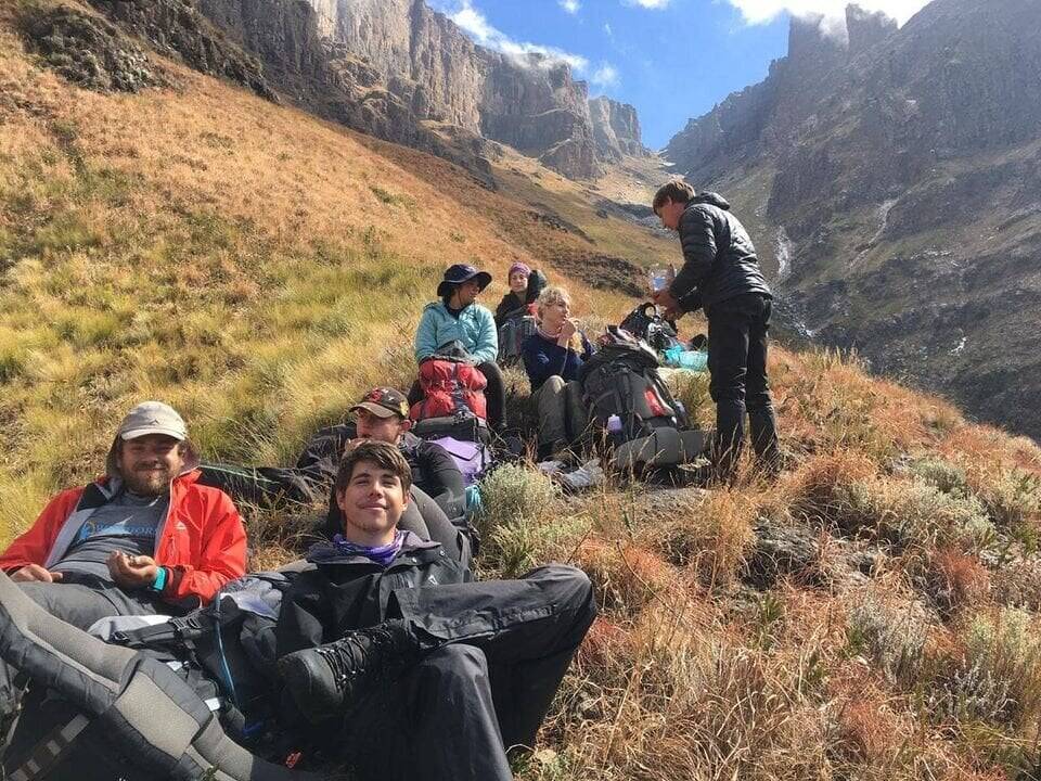A group of people with backpacks lounge on a hill with large mountains in the background.