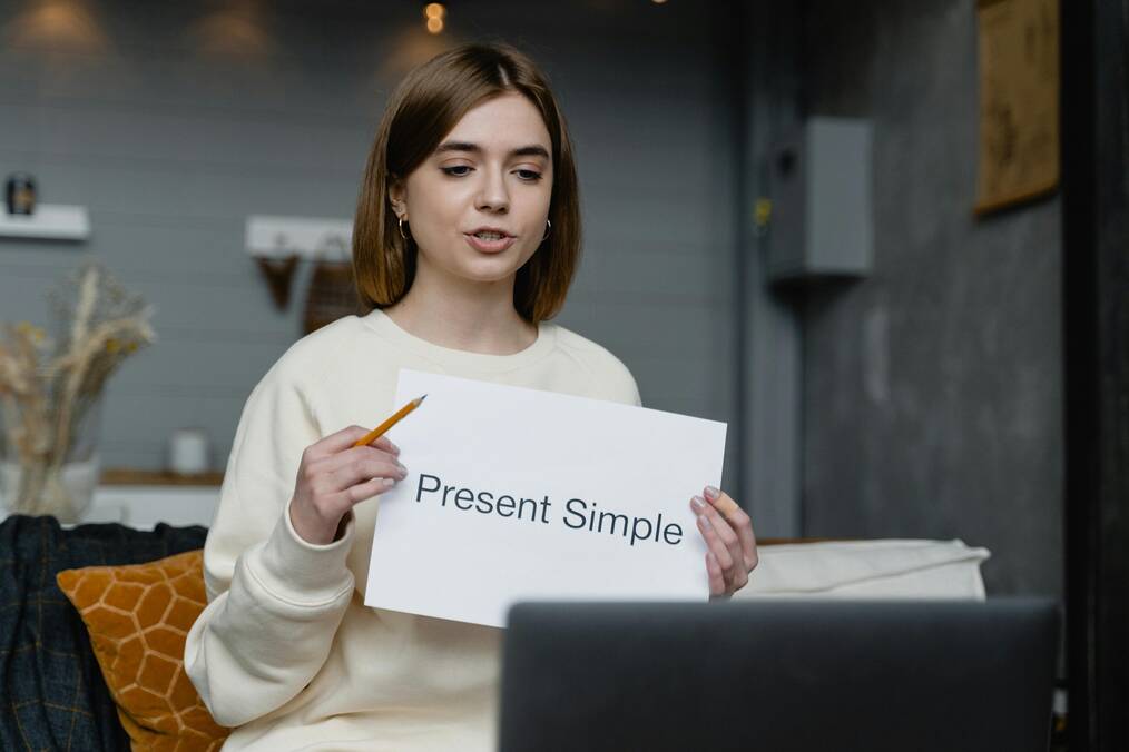 A woman holds a paper up to a computer screen that says, "present simple" as she teaches online.