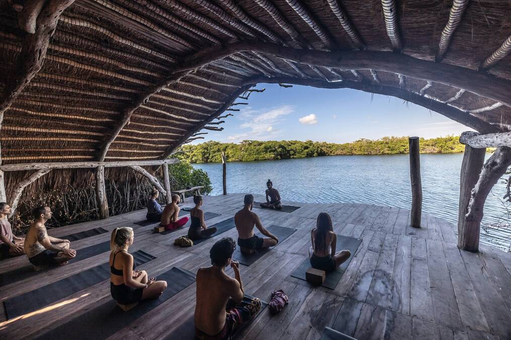 A group of people sit on yoga mats under a wooden structure looking out at a lake.