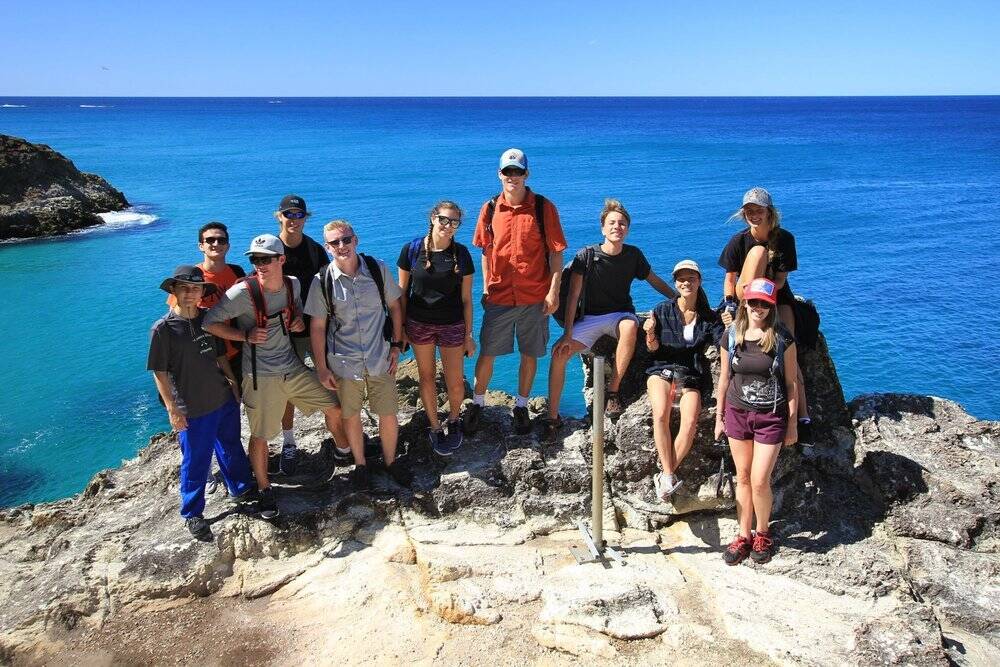 A group of young adults wearing backpacks pose for a photo on a cliff near a blue ocean.