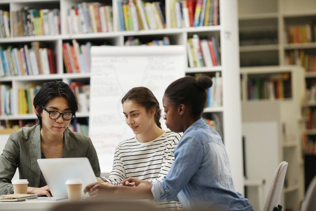 Three students sitting at table studying inside library in front of row of books
