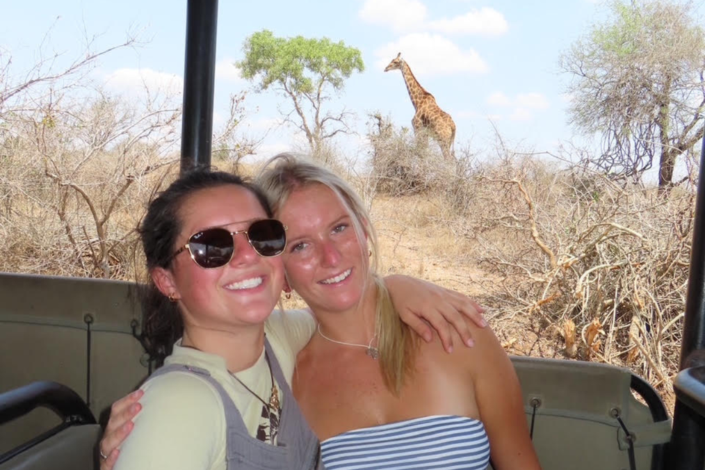 Two students smiling on a safari, in front of a giraffe