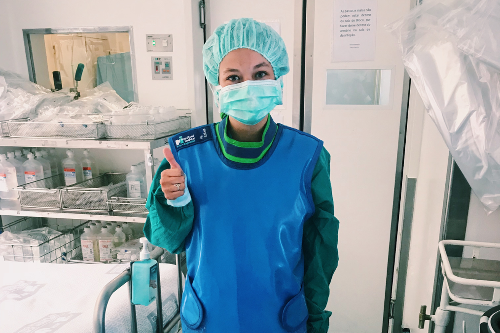A pre-health shadowing student on an Atlantis internship abroad gives a thumbs up in scrubs, a surgical cap, and a mask inside a hospital operating room