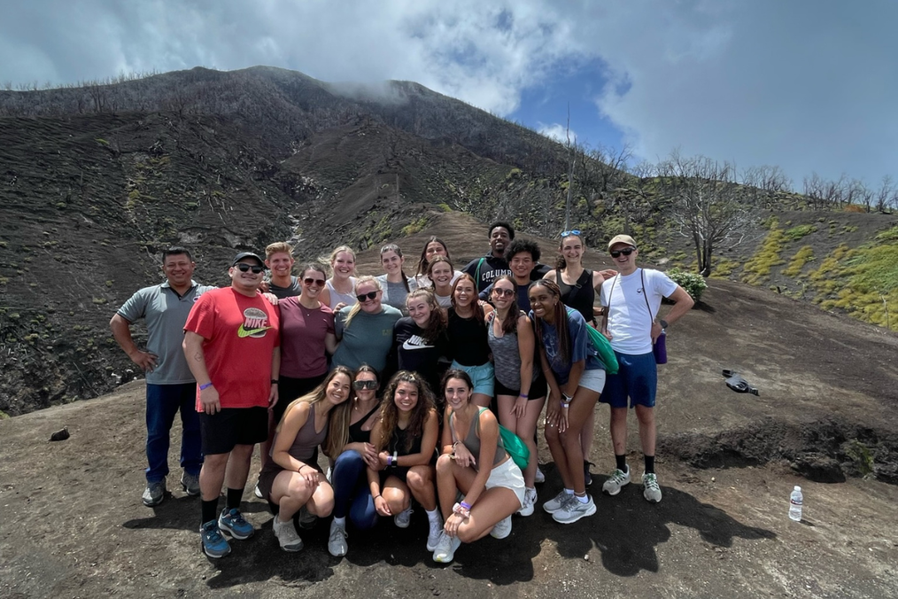 A large group of Toucan Abroad participants poses together on an ash-covered volcanic hillside in Costa Rica, with a cloud-covered peak rising behind them
