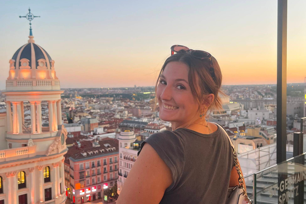A woman looks back over her shoulder on a Madrid rooftop terrace at sunset during her time with The Intern Group's Madrid program, with the domed Círculo de Bellas Artes building and city skyline behind her