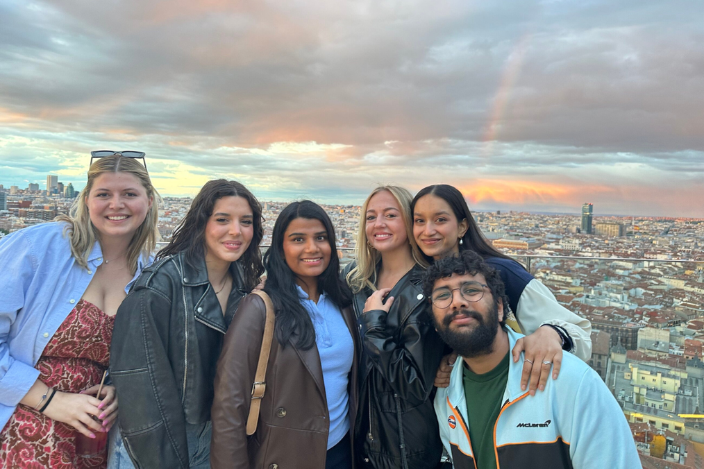 Six friends from The Intern Group's Madrid program pose together on a rooftop at sunset, with a rainbow arcing over the Madrid skyline behind them