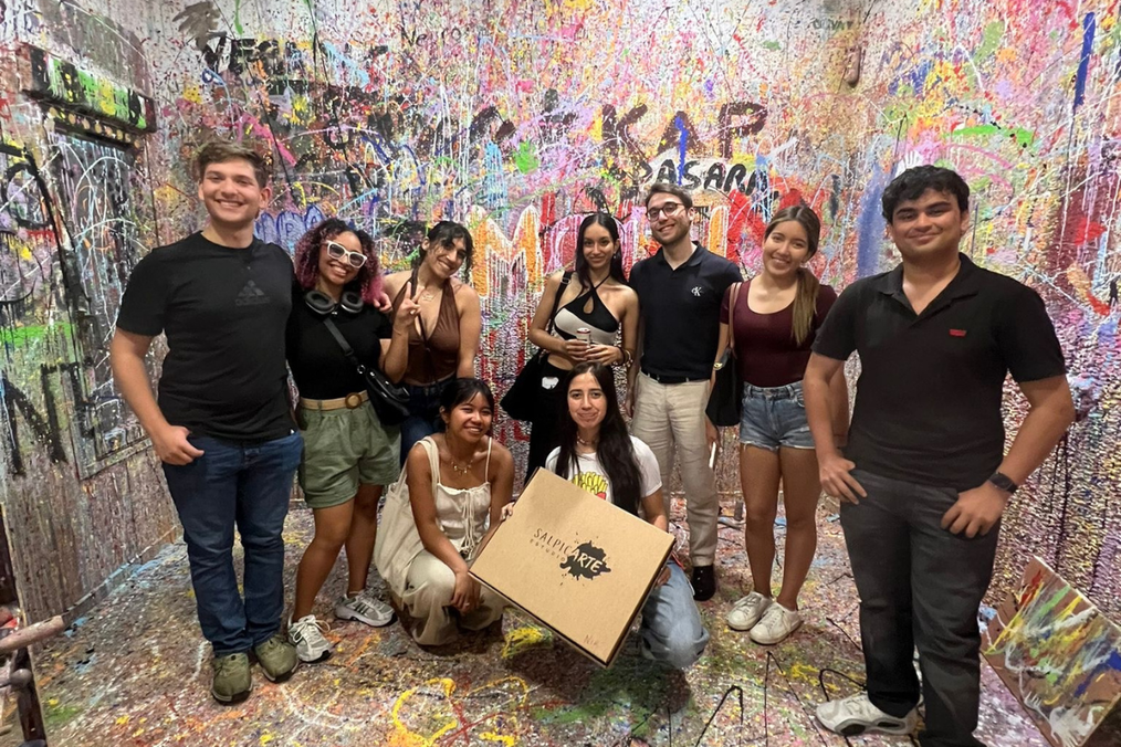 Nine friends from The Intern Group's Madrid program pose together in a paint-splattered room at SalpicArte Estudio, one holding a branded box, with graffiti-covered walls behind them