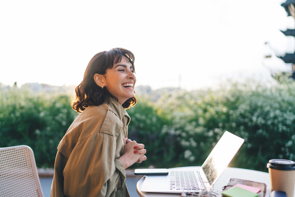 A woman laughs while sitting at an outdoor table with her laptop and a coffee, surrounded by greenery