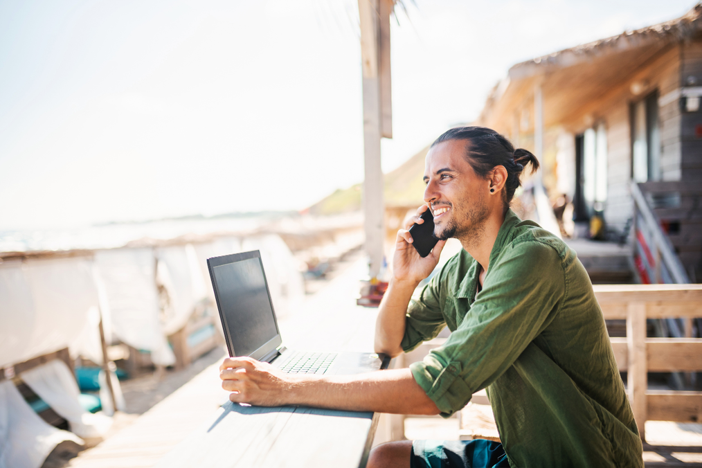 A man smiles while talking on his phone with his laptop open on a wooden deck overlooking a beach