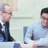 A male professor sits at a table next to a male student