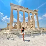 A young woman with long brown hair, wearing a black tank top and light denim shorts with a straw hat, stands in front of temple ruins with columns still standing.