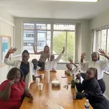 A group of people gathered around a table wave at the camera