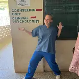 A volunteer in scrubs poses happily in front of a sign that reads 'Counselling Unit' and 'Clinical Psychologist'