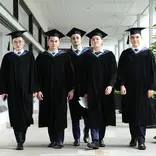 Five graduates in caps and gowns walk together down a hallway, facing forward and looking at the camera.