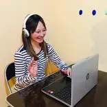 A Japanese teacher conducting an online 1-on-1 lesson with a student via Zoom. The teacher is smiling and explaining a lesson in Japanese using teaching materials on the screen.