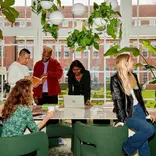 A group of students standing around a long table in front of a large white-pane window. The students are looking through text books, working on laptops, drinking coffee and chatting and laughing with one another. 