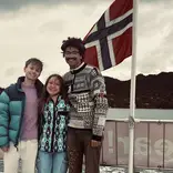 three college students stand smiling with the Norwegian flag waving in the wind