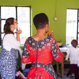 A volunteer teacher and a local student stand before a classroom of young children in Tanzania. They are pointing to their mouths to demonstrate pronunciation against a backdrop of bright green walls.