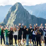 A group of teenagers standing on a stone terrace at Machu Picchu, waving their hands in the air with the ancient Incan ruins and Huayna Picchu mountain in the background.