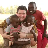 Volunteer kneeling outdoors with three young children, smiling and embracing them during a community program at Maisha.