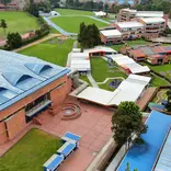The image shows an aerial view of an educational institution with a spacious and well-organized campus, featuring brick buildings with blue and white roofs, well-maintained green areas, recreational spaces, and sports facilities such as an athletic track. Walkways and courtyards connect the different areas, and the surroundings combine nature with urban buildings, conveying a sense of order, openness, and institutional well-being.