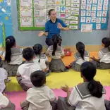 TEFL teacher sitting at the front of a classroom, engaging a group of young students in a lesson with hand gestures and visual aids on the wall.