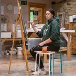 Woman sitting on stool in front of easel with paintbrush