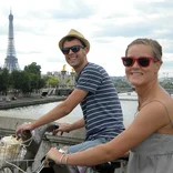 Two students wearing sunglasses riding bikes across the Seine River with the Eiffel Tower behind them