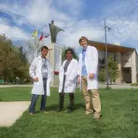 three students wearing lab coats standing on a grassy lawn in front of a building