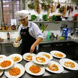 student in a commercial kitchen, wearing a chefs hat, sprinkling cheese on multiple plates of pasta