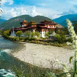 A large traditional Bhutanese fortress-monastery sits beside a winding river, surrounded by forested mountains under a bright, partly cloudy sky.