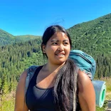 A brown-skinned Asian woman with a teal hiking backpack stops on the trail and poses in front of a lush view of the mountains