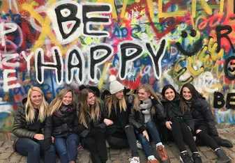 seven girls sitting in front of the John Lennon Wall with the prominent phrase "BE HAPPY" behind them
