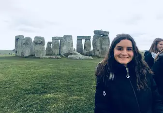 This photo shows a girl standing in front of Stonehenge. She is smiling and wearing a black coat, and it's cloudy behind her. The grass around Stonehenge is very green and contrasts nicely with the gray of the rocks. 