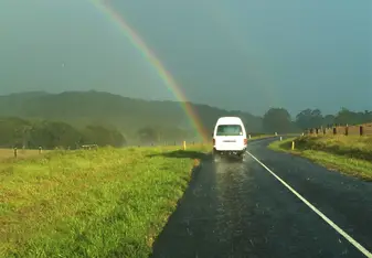 A white van is driving down a local road. There is a rainbow coming from the left to meet the van.