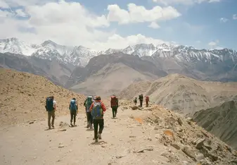 A landscape with snow-capped mountains in the background. Blue sky with clouds. Several hikers with backpacks walking away from the camera on a dirt trail.