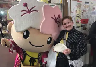 A Japanese Mascot with plum and sakura blossoms on its' head next to a person wearing a yellow tie and holding a pierogi 