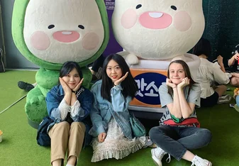 3 girls sitting on the ground in front of characters doing the korean flower pose