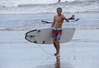 Teen boy gives thumbs up with surf board by waves