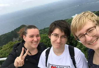 The image is a horizontal picture of three girls from the chest up, looking at the camera. One makes a peace sign and smiles, another grimaces at the camera, and the last smiles at the camera as well. Behind them is a landscape with lush greenery, behind that the ocean, and a cloudy sky above them.