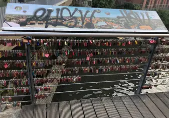 A picture of a bridge above a river. There's a panoramic picture of the town of Luneburg that's been graffitied over. On the bridge, there are hundreds of love locks hanging off of suspension wire. 