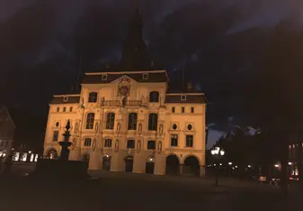 A picture taken at night of an imposing building lit up from below, casting the building in an orange hue. This is the town hall, it has several balconies and a copper roof. There's some marble statues on the facade of the building and the silhouette of a fountain in the foreground. 