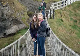 Two young women are standing in the middle of Carrick-a-Rede rope bridge, smiling at the camera with a green mountain view in the background.