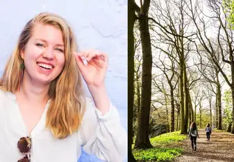 There is a smiling young woman moving her hair behind her ear, standing in front of a bright blue painted wall. Beside her, there is a picture of two people walking on a footpath between very tall, green trees.