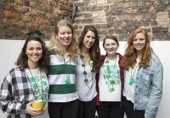 Five young women are smiling, wearing green beaded necklaces and face paint celebrating St. Patrick's Day.